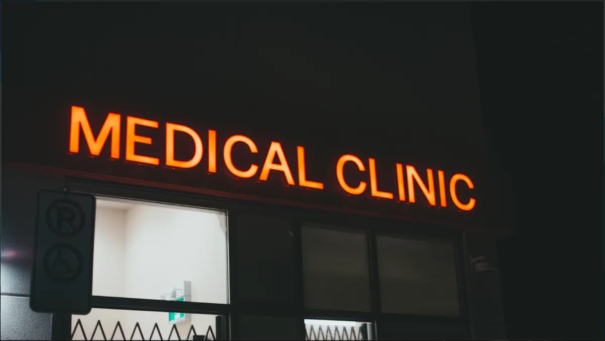 A modern clinic consultation room with a doctor reviewing a bilingual SOAP note on a large monitor, an AI Clinical Assistant side-panel suggesting ICD-10 codes, and a tablet on the desk showing an Arabic patient summary.